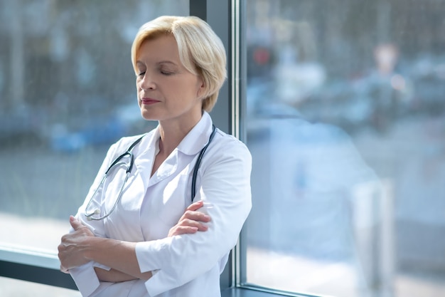 Medical profession. Tired female doctor folding arms, standing next to the window with closed eyes