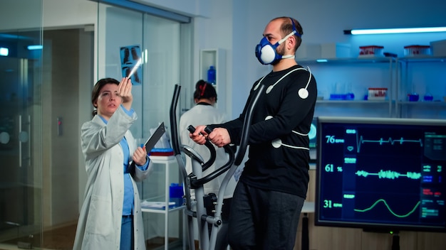 Ricercatore medico che esamina l'evoluzione delle condizioni di salute dell'atleta, guardando i raggi x mentre l'uomo con la maschera corre su cross trainer monitorando la sua resistenza