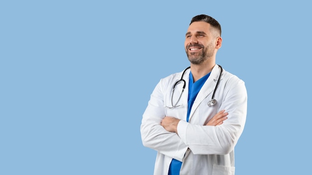 Medical services concept Happy male doctor in uniform posing with folded arms over blue background panorama free space