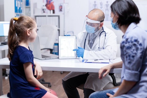 Medical specialist presenting skeleton using tablet sitting on desk in medical office