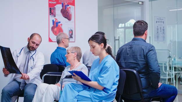 Medical staff discussing diagnosis with disabled senior woman in wheelchair in hospital hallway. Old man waiting for medical examination. Nurse taking notes on clipboard.