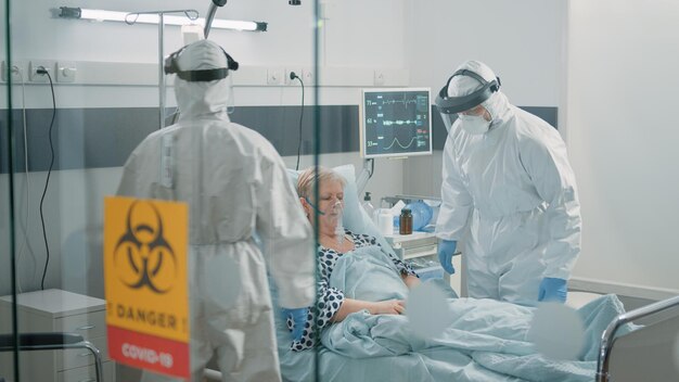 Il personale medico in tute ignifughe si occupa della consulenza di pazienti anziani con coronavirus. donna malata in quarantena del reparto ospedaliero riceve visita di controllo da paramedici con uniformi e maschere protettive