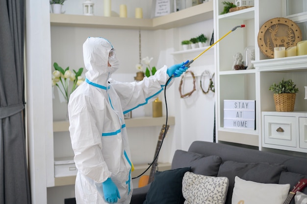 A medical staff in ppe suit is using disinfectant spray in living room