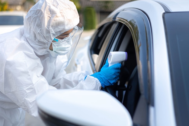 Medical staff with PPE use infrared thermometer gun to check body temperature at drive thru station in hospital. New normal healthcare and medical concept.