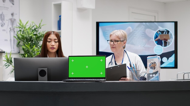 Medical staff working at reception desk with greenscreen\
display on laptop, using blank copyspace screen. hospital lobby\
with waiting area, isolated chroma key template and mockup\
background.