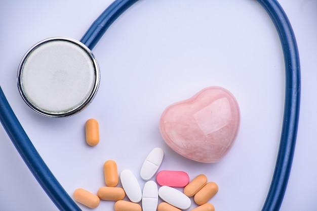 Medical stethoscope, pills and pink glass heart on white surface. Cardiology concept