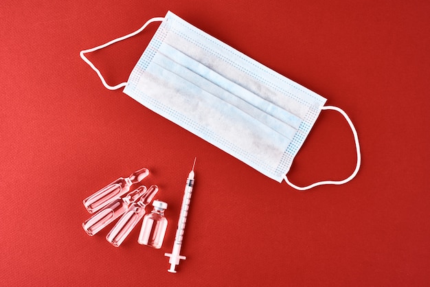 Medical syringe, ampoule with medicine and protect mask on red background, top view. Vaccination and epidemic virus protection concept.