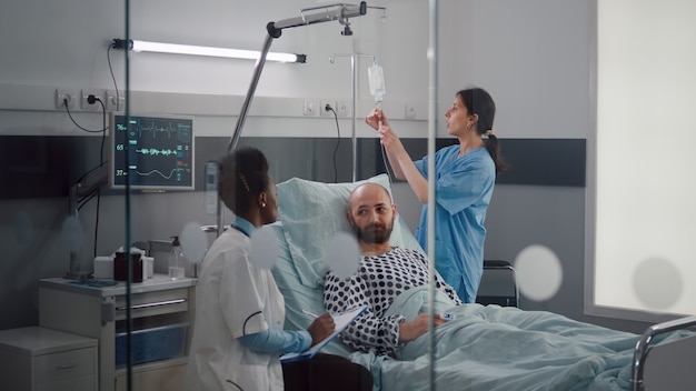 Medical team monitoring patient vital checking heart rate injecting fluids in drip bag for remission therapy in hospital ward