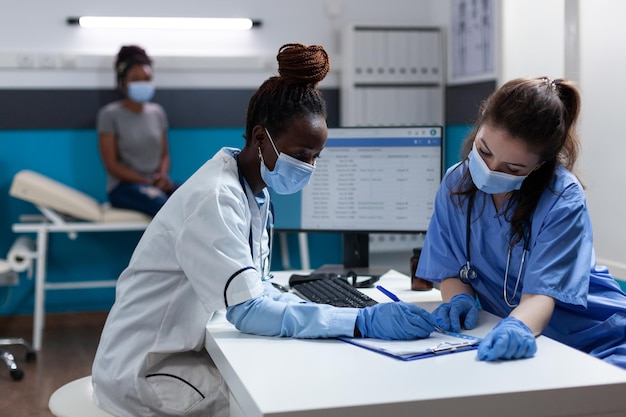 Medical team with protection face mask to prevent infection with covid19 analyzing consultation documents working in hospital office. African american doctor examining sick patient during appointment