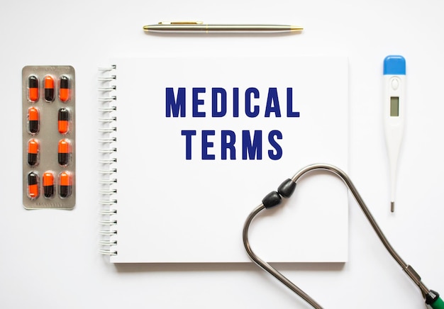 MEDICAL TERMS is written in a notebook on a white table next to pills and a stethoscope