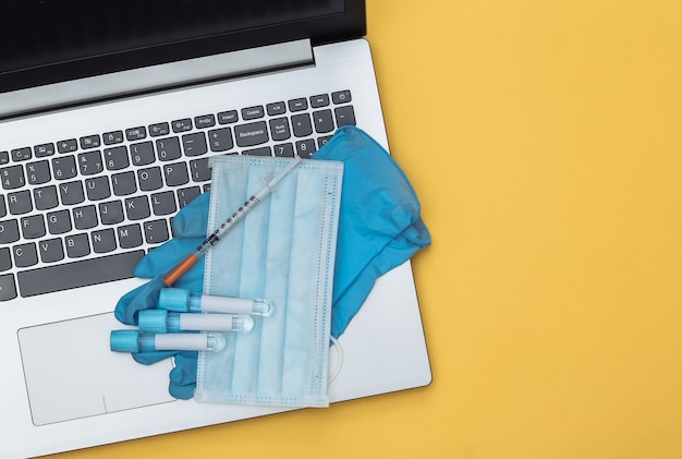 Medical test tubes, face mask, gloves with syringe on  laptop keyboard. Yellow Background. Top view