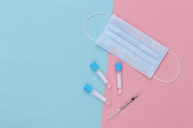 Medical test tubes with syringe and face mask on blue pink pastel background. Vaccination. Top view