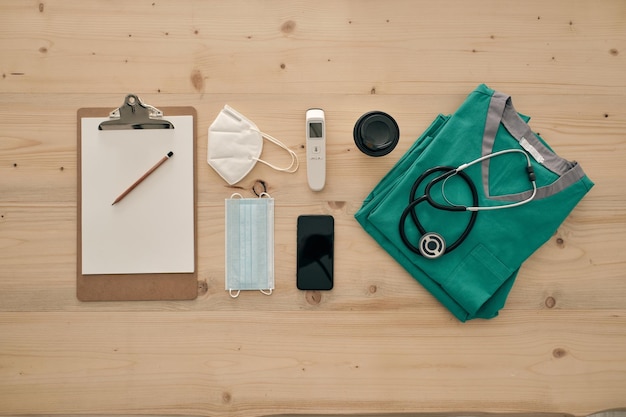 Medical uniform with stethoscope near cellphone and respiratory masks with coffee to go and blank paper sheet on desk