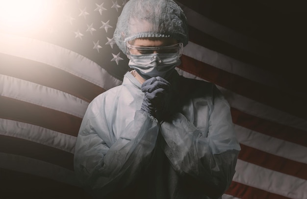 Medical worker in a gown with gloves and a medical mask is praying against the background of the American flag