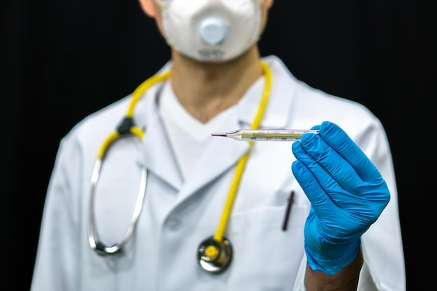 A medical worker holds a medical mercury thermometer in his hands Measurement of body temperature during coronavirus infection A doctor in a medical gown and mask on a black background