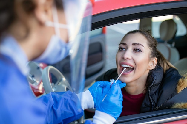 Medical worker performing drivethru COVID19 checktaking nasal swab specimen sample from female patient through car windowPCR diagnostic for Coronavirus presencedoctor in PPE holding test kit