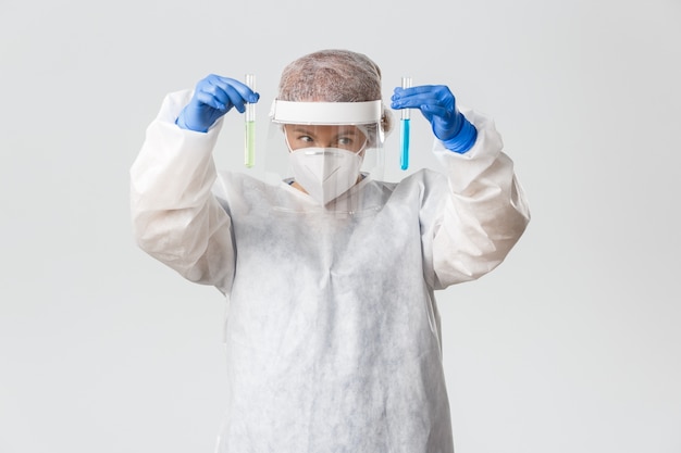 Medical workers, covid-19 pandemic, coronavirus concept. Cheerful smiling female doctor in personal protective equipment comparing two test-tubes with samples of vaccine, grey background.