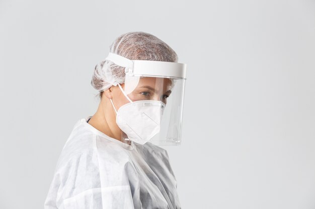 Medical workers, covid-19 pandemic, coronavirus concept. Profile of serious-looking female doctor in personal protective equipment, face shield and respirator listening to patient, provide checkup.