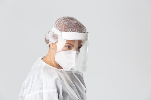 Medical workers, covid-19 pandemic, coronavirus concept. Profile of surprised female doctor in personal protective equipment looking right lower angle at patient, standing grey background.