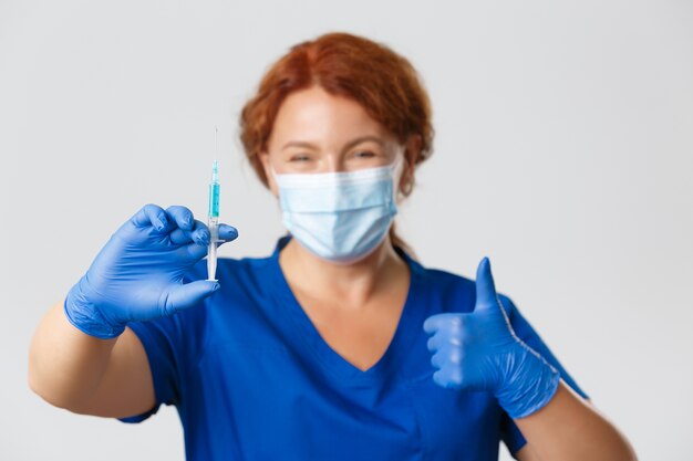 Medical workers, covid-19 pandemic, coronavirus concept. Smiling female doctor, physician in face mask, rubber gloves prepare syringe with vaccine for injection, making flu shot, thumbs-up.