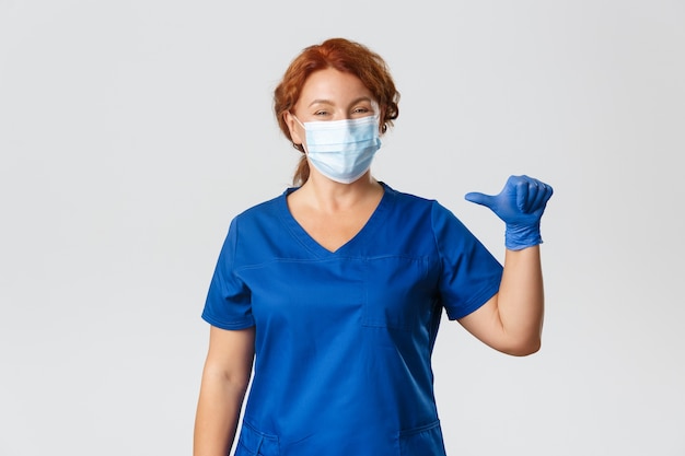 Medical workers, covid-19 pandemic, coronavirus concept. Smiling pleasant nurse, female professional doctor in medical mask, gloves and scrubs pointing at herself, being real pro, grey background.