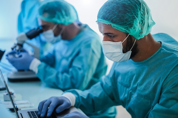 Foto operatori sanitari in tuta ignifuga che lavorano con computer portatile e micoroscopio all'interno dell'ospedale di laboratorio durante l'epidemia di coronavirus