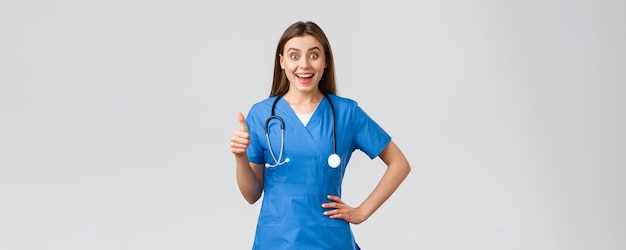 Medical workers healthcare covid19 and vaccination concept Surprised excited female nurse or doctor in blue scrubs with stethoscope show thumbsup in approval like idea smiling upbeat