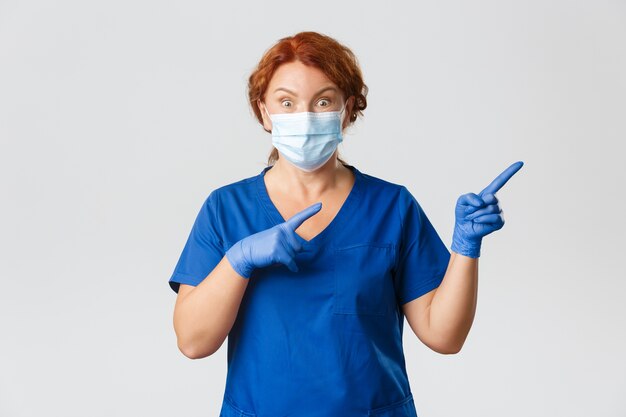 Medical workers,  pandemic, coronavirus concept. Surprised and impressed female doctor, middle-aged physician in face mask, gloves pointing upper right corner with amazed expression