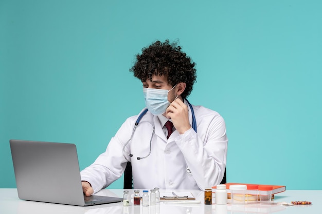 マスクを身に着けている白衣でリモートでコンピューターに取り組んでいる医療の若いハンサムな医師