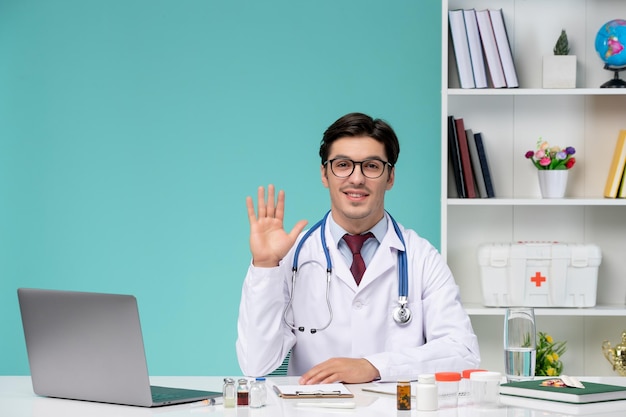 Medical young smart doctor in lab coat working remotely on computer saying hello to patient