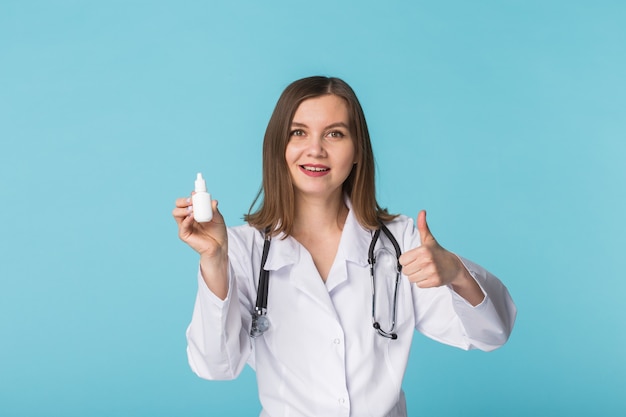 Medicaments and pharmaceutical concept - Young female doctor or pharmacist holding nasal spray with thumbs up on blue background