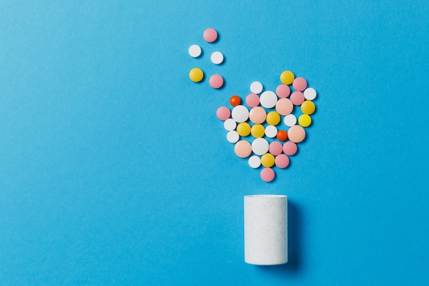 青い背景に分離されたハートの形で薬の白、カラフルな丸い錠剤。丸薬の幾何学的形状、ボトル。健康、治療、選択、健康的なライフスタイルの概念。宇宙広告をコピーします。