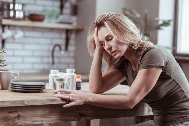 Medicijnen nemen. Blondharige vrouw zit in de keuken en neemt medicijnen terwijl ze zich ziek en moe voelt