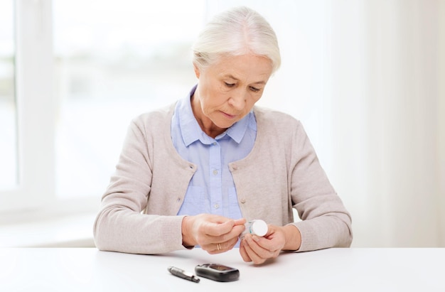 medicine, age, diabetes, health care and people concept - senior woman with glucometer and test stripes checking blood sugar level at home