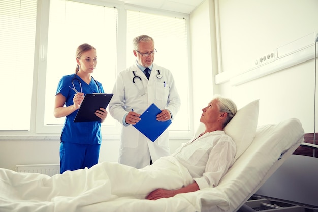 medicine, age, health care and people concept - doctor and nurse with clipboards visiting senior patient woman at hospital ward