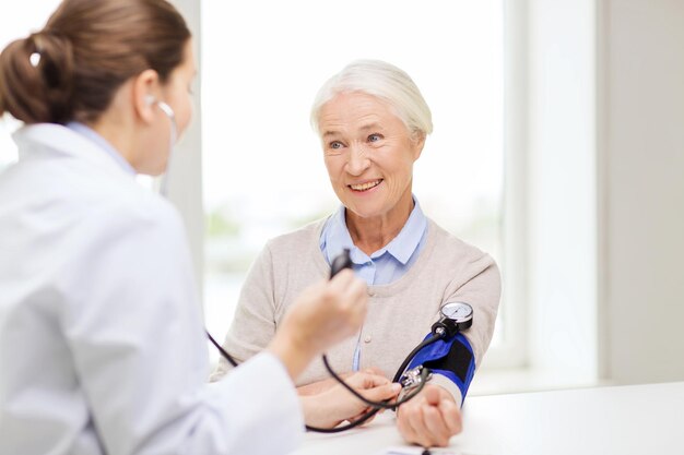 medicine, age, health care and people concept - doctor with tonometer checking happy senior woman blood pressure level at hospital