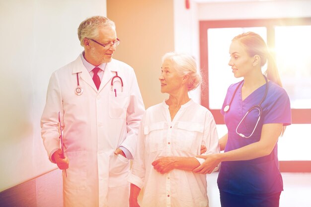 medicine, age, health care and people concept - male doctor with clipboard, young nurse and senior woman patient talking at hospital corridor