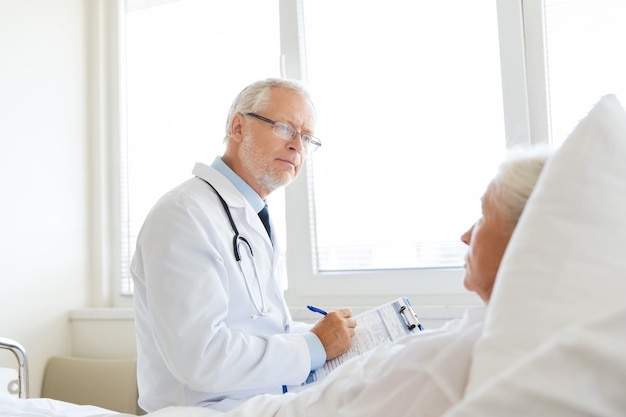 medicine, age, health care and people concept - senior woman and doctor writing to clipboard at hospital ward