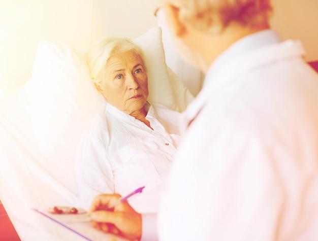 medicine, age, health care and people concept - senior woman and doctor writing to clipboard at hospital ward