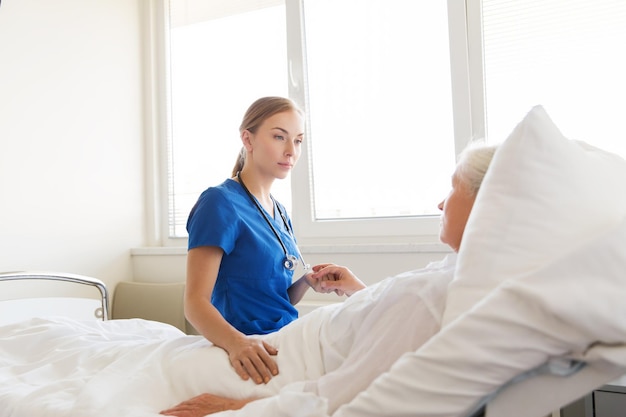 medicine, age, support, health care and people concept - doctor or nurse visiting and cheering senior woman lying in bed at hospital ward