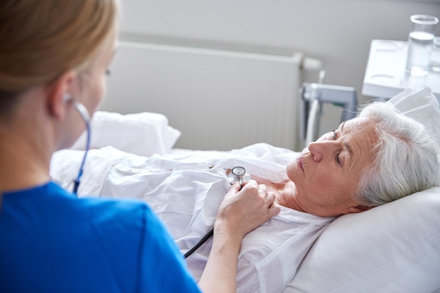 Photo medicine, age, support, health care and people concept - doctor or nurse with stethoscope visiting senior woman and checking her breath or heartbeat at hospital ward
