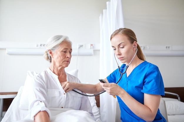 medicine, age, support, health care and people concept - doctor or nurse with stethoscope visiting senior woman and checking her heartbeat at hospital ward