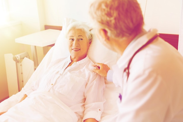 medicine, age, support, health care and people concept - doctor visiting and cheering senior woman lying in bed at hospital ward