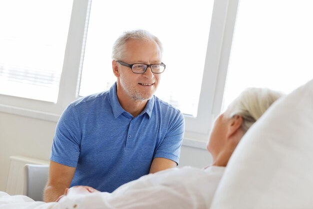 medicine, age, support, health care and people concept - happy senior man visiting and cheering his woman lying in bed at hospital ward