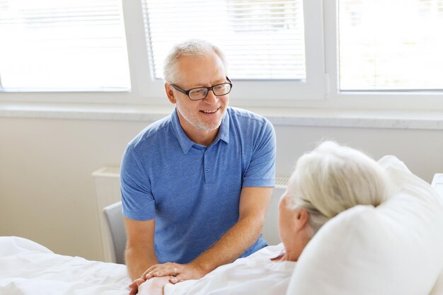 medicine, age, support, health care and people concept - happy senior man visiting and cheering his woman lying in bed at hospital ward