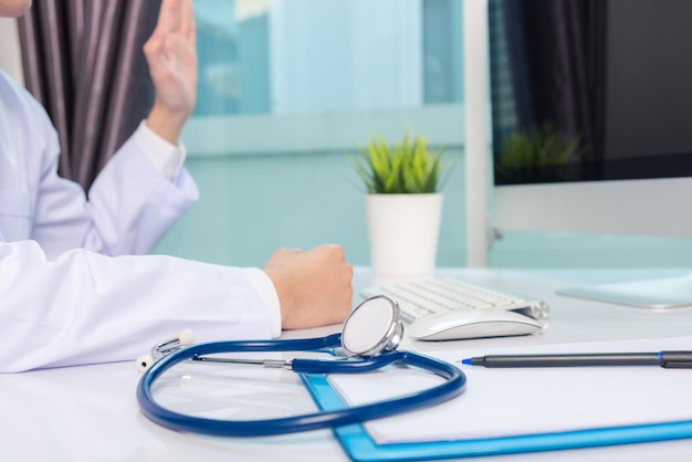 Medicine doctor's working on desk. Closeup of Stethoscope. Hand of man physician video call raise hands to greet patients on table front computer monitor at hospital office, Healthcare medic concept