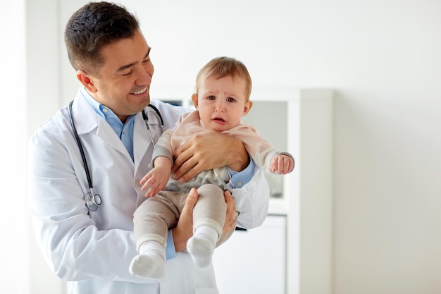 medicine, healtcare, pediatry and people concept - happy doctor or pediatrician holding sad crying baby girl on medical exam at clinic