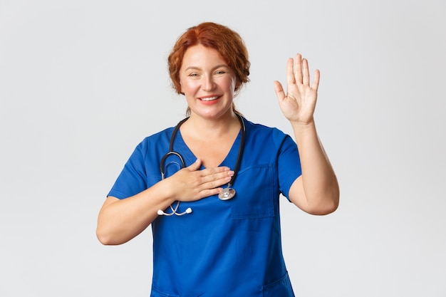 医学、ヘルスケアの概念。スクラブでキュートで楽観的な中年の女性医師、看護師、または医療従事者の笑顔は、片手を上げて手のひらを心臓に置き、誓い、約束をします。