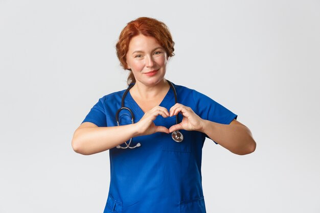 Medicine, healthcare and coronavirus concept. Caring middle-aged female nurse, doctor in scrubs showing heart gesture and smiling, looking after patients at retirement home, grey background.