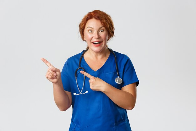 Medicine, healthcare and coronavirus concept. Excited cheerful redhead nurse, medical worker in scrubs pointing upper left corner. Doctor showing awesome promo offer, smiling amazed.